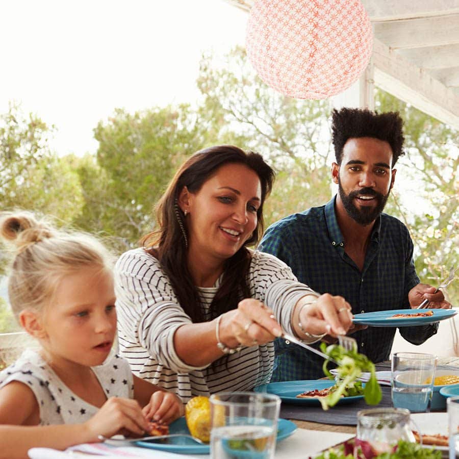 Three people, two adults and a child, are sitting at a table outdoors, enjoying a meal together. The woman is serving salad, while the others are smiling. Trees and a pink lantern are visible in the background.